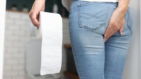 Frau steht in einem Badezimmer, trägt eine Jeans und und ein weißes Oberteil. In ihrer linken Hand hält sie eine Klorolle, mit der Rechten fasst sich sich an das Hinterteil. Im Hintergrund ist die Toilettenschüssel. (Foto: Colourbox, 48158578)