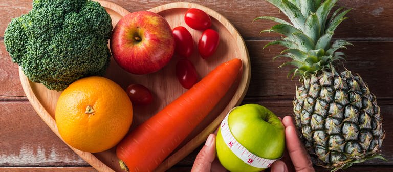 Brokkoli, Tomaten, Orange, Apfel, Karotte liegen auf einem Holzteller, daneben Ananas und Apfel. Hohe Cholesterinwerte sind ein Risikofaktor für Herzinfarkt und Schlaganfall - gefährlicher als HDL ist der LDL-Wert. (Foto: Colourbox, 56940209)
