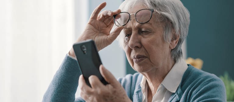 Eine ältere Frau schiebt mit ihrer rechten Hand ihre Brille auf die Stirn. In ihrer linken Hand hält sie ein Handy. (Foto: Adobe Stock, PhotoPro)