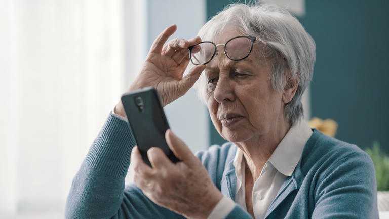Eine ältere Frau schiebt mit ihrer rechten Hand ihre Brille auf die Stirn. In ihrer linken Hand hält sie ein Handy. (Foto: Adobe Stock, PhotoPro)