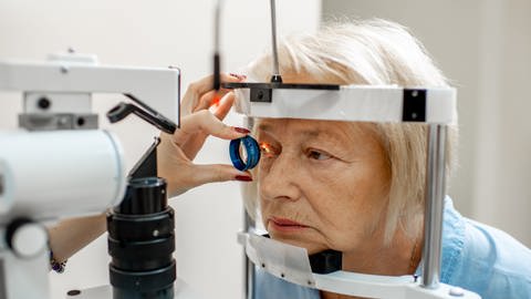Eine Augenärztin hält mit ihrer linken Hand eine Linse vor das rechte Auge der Patientin. Ein Lichtstrahl fällt auf das Auge. Ihr Gesicht steckt in einem Messgerät. (Foto: Adobe Stock, rh2010)