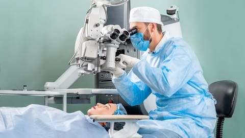 Ein Augenarzt in Operationskleidung sitzt an einem großen Mikroskop und schaut sich die Augen einer Patientin an. Er bereitet sich auf eine Laser-Operation der Augen  vor. (Foto: Adobe Stock)
