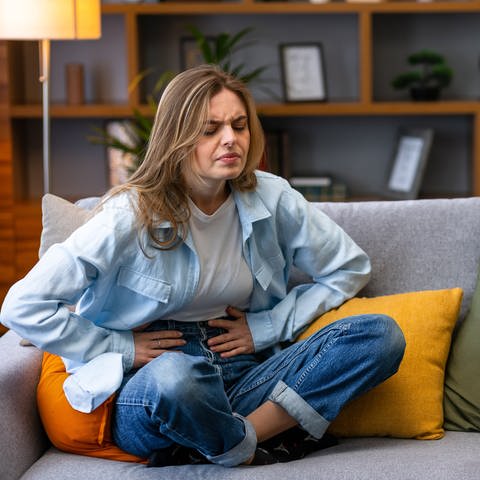 Eine Frau sitzt mit Bauchschmerzen auf einem Sofa und hält ihren Bauch mit den Händen. (Foto: Adobe Stock, Adobe Stock/Ivan)