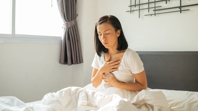 Frau in weißem T-Shirt sitzt im Bett und hält sich Brust und Magen. Was hilft wirklich gegen Sodbrennen und Reflux? Wann ist eine Operation notwendig? Welche Nebenwirkungen haben Säurehemmer? (Foto: Adobe Stock, Adobe Stock | sutlafk)