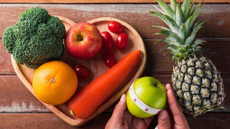 Brokkoli, Tomaten, Orange, Apfel, Karotte liegen auf einem Holzteller, daneben Ananas und Apfel. Hohe Cholesterinwerte sind ein Risikofaktor für Herzinfarkt und Schlaganfall - gefährlicher als HDL ist der LDL-Wert. (Foto: Colourbox, 56940209)