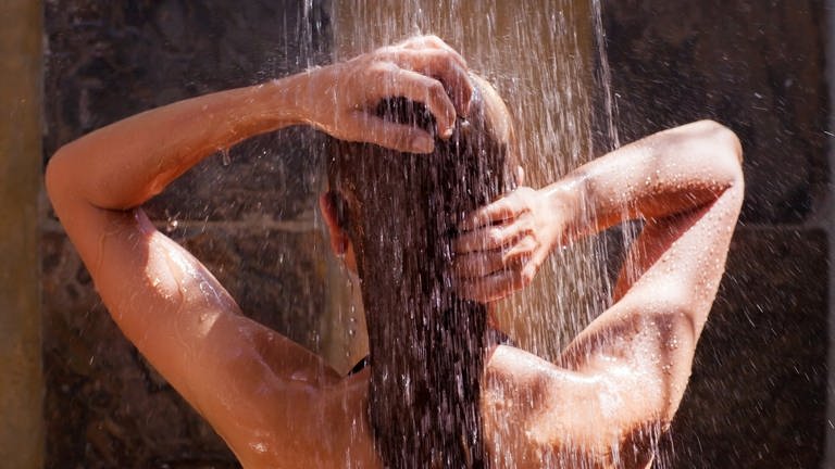 Eine Frau steht unter der Dusche und fährt sich über die Haare. Sie ist von hinten zu sehen.