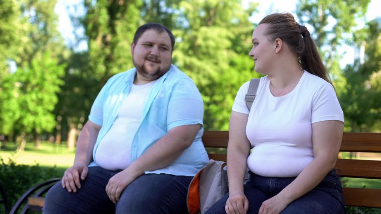 Übergewichtige Frau und übergewichtiger Mann sitzen in der Natur auf einer Bank und schauen sich gegenseitig in die Augen. Beide lächeln sich an.