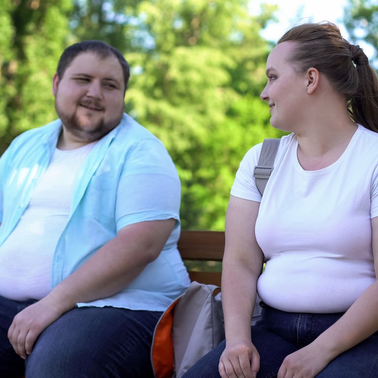 Übergewichtige Frau und übergewichtiger Mann sitzen in der Natur auf einer Bank und schauen sich gegenseitig in die Augen. Beide lächeln sich an.