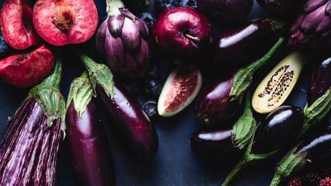 Verschiedene violette Obst- und Gemüsesorten, wie Auberginen, Artichocken, Feigen und Pflaumen liegen auf dunkler Schieferplatte. Bitterstoffe sind gesund!
