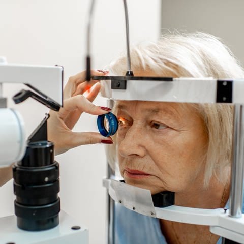 Eine Augenärztin hält mit ihrer linken Hand eine Linse vor das rechte Auge der Patientin. Ein Lichtstrahl fällt auf das Auge. Ihr Gesicht steckt in einem Messgerät.