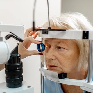 Eine Augenärztin hält mit ihrer linken Hand eine Linse vor das rechte Auge der Patientin. Ein Lichtstrahl fällt auf das Auge. Ihr Gesicht steckt in einem Messgerät. (Foto: Adobe Stock, rh2010)
