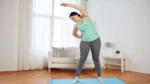 Übergewichtige Frau in Sportkleidung steht in ihrem Wohnzimmer barfuß auf einer Sportmatte. Ihr rechter Arm ist in die Hüfte gestemmt, ihr linker Arm über den Kopf gestreckt.