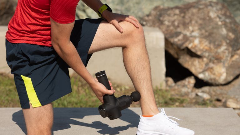 Ein Jogger hält sich eine Massagepistole an den Unterschenkel. Er trägt Sportklamotten und es scheint die Sonne. (Foto: Adobe Stock, AdobeStock / lashkhidzetim)