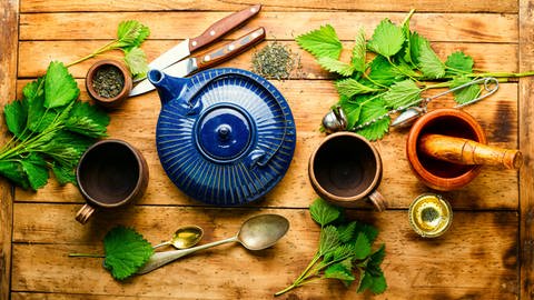Auf einem Tisch liegen Brennnesselblätter, Löffel, eine Teekanne, ein Mörser und Tee-Eier. Brennnesselblätter können zum Beispiel als Tee zubereitet werden.