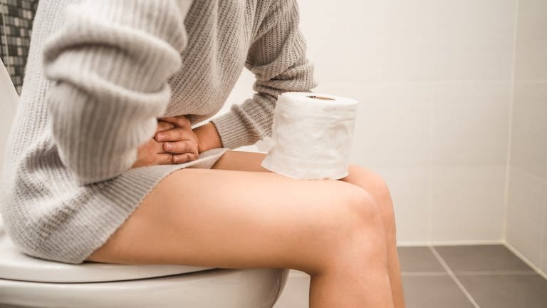 Was tun bei chronischer Verstopfung? Eine Person sitzt auf der Toilette und hält sich den Unterbauch. Auf ihren Beinen steht eine Rolle Toilettenpapier. (Foto: Adobe Stock, AdobeStock / Sirichai)