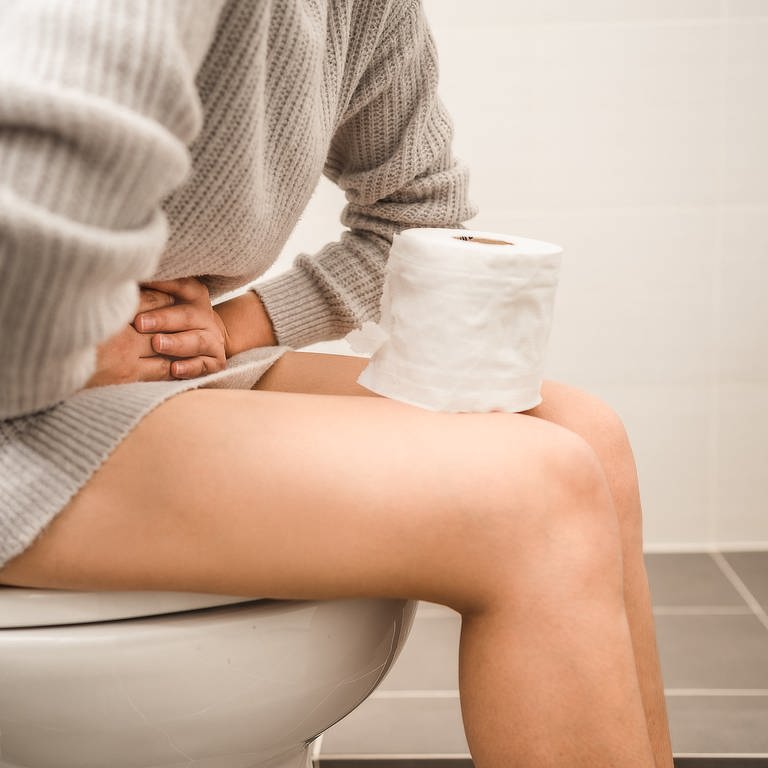 Was tun bei chronischer Verstopfung? Eine Person sitzt auf der Toilette und hält sich den Unterbauch. Auf ihren Beinen steht eine Rolle Toilettenpapier.