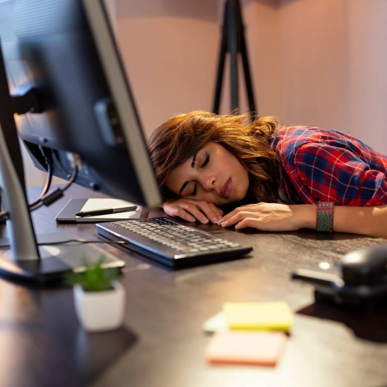 Junge Frau schläft vor ihrem Computer mit dem Kopf auf dem Tisch. Was bringen Mittagsschlaf und Powernap gegen Müdigkeit? (Foto: Adobe Stock)