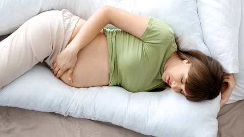 Eine schwangere Frau schläft auf der Seite. Ein U-förmiges Kissen stützt ihren Kopf und ihren Rücken, sodass sie nicht in die gefährliche Rückenlage kommt. (Foto: Adobe Stock)