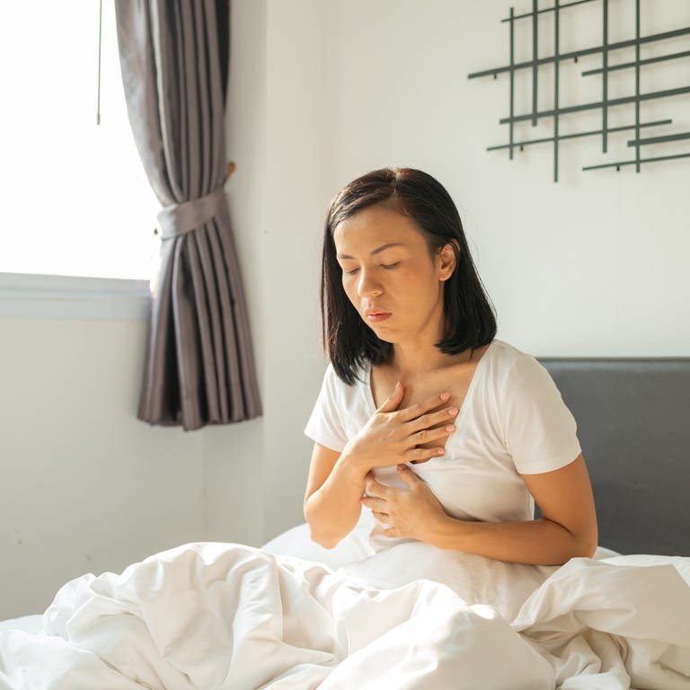 Frau in weißem T-Shirt sitzt im Bett und hält sich Brust und Magen. Was hilft wirklich gegen Sodbrennen und Reflux? Wann ist eine Operation notwendig? Welche Nebenwirkungen haben Säurehemmer?