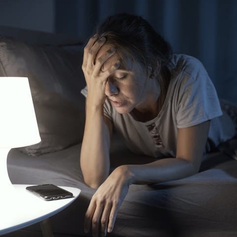 Frau liegt auf dem Bauch im Bett und hält sich schmerzverzerrt das Gesicht. Chronische Schmerzen: Wie entsteht ein Schmerzgedächtnis? Welche Therapie hilft?