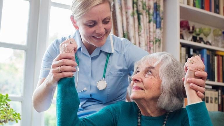 Eine Seniorin wird von einer Pflegerin beim Aufstehen gestützt. (Foto: getty images -)