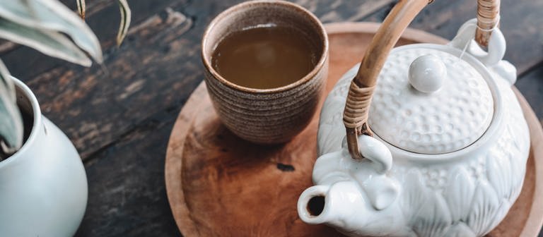Braune Tasse mit Tee und eine weiße Kanne mit Henkel auf einer braunen Platte. Links sieht man eine Pflanze in einem weißen Topf. Die Gegenstände liegen auf einem dunkelbraunen Tisch aus Holz (Foto: Unsplash / Content Pixie)