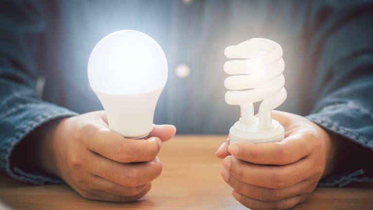 Eine Person sitzt an einem Holztisch und hält jeweils eine LED-Lampe in der Hand, die leuchten. (Foto: Adobe Stock, Kt Stock)