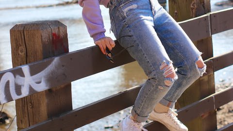 Mädchen mit zerschlissenen Jeans sitzt auf einem Zaun. Jeans im Used Look schaden in der Herstellung der Umwelt. Geht das auch nachhaltig? (Foto: unsplash/Spencer Quast)