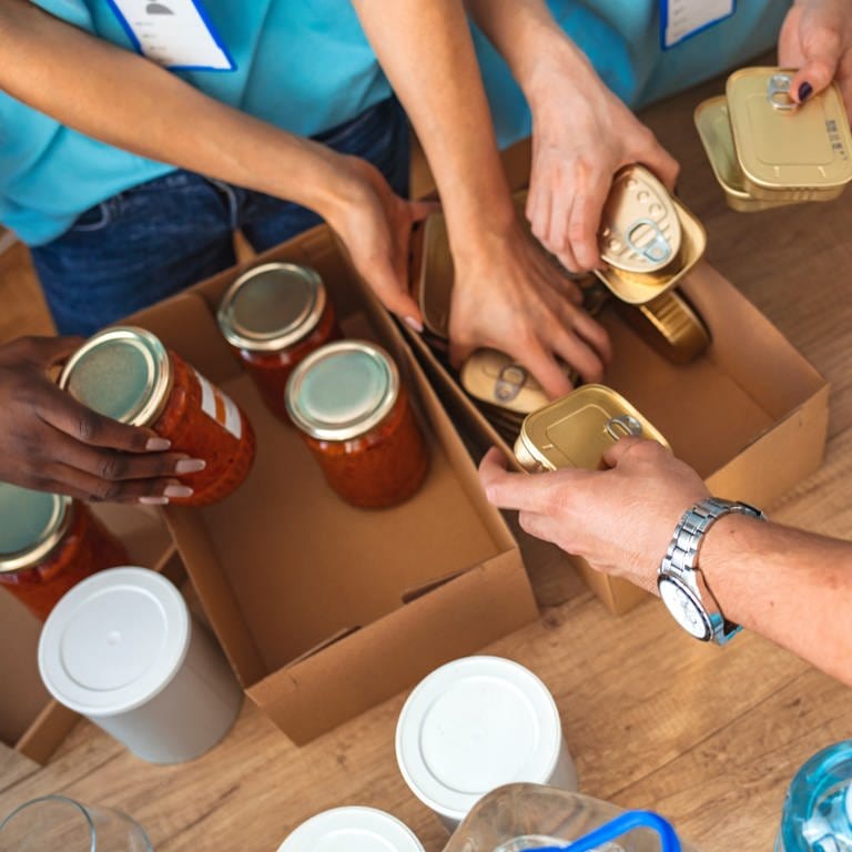 Mehrere Hände Hände greifen nach mit Tomatensoße gefüllten Konservengläsern und goldenen Sardellen-Blechdosen, die in zwei Kartons gestapelt sind. (Foto: SWR)