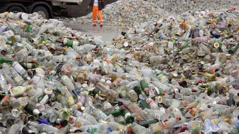 Es stapeln sich bunte Altglasflaschen in rießigen Haufen im Hof einer Sortieranlage. Im Hintergrund ist ein Mitarbeiter zu erkennen. (Foto: dpa Bildfunk, Picture Alliance)