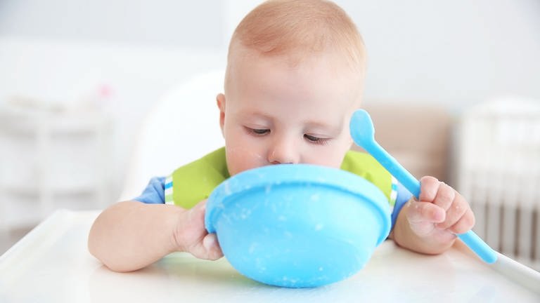 Ein kleines Kind sitzt an einem Tisch und hält Kindergeschirr, eine blaue Schüssel und einen Löffel, in der Hand. Das Geschirr ist aus Melaminharz. (Foto: Adobe Stock)