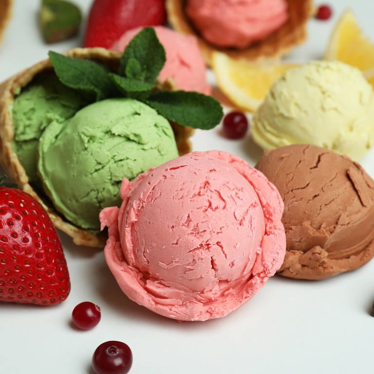 Verschiedene Eissorten liegen kugelweise mit frischem Obst auf einem weißen Untergrund.