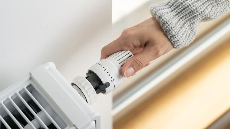 Eine Person mit grauem Pulli greift von rechts das Thermostat einer Heizung, die auf Stufe 2 gestellt ist.