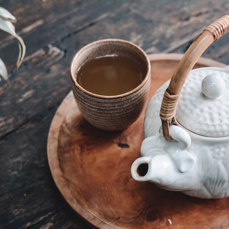 Braune Tasse mit Tee und eine weiße Kanne mit Henkel auf einer braunen Platte. Links sieht man eine Pflanze in einem weißen Topf. Die Gegenstände liegen auf einem dunkelbraunen Tisch aus Holz