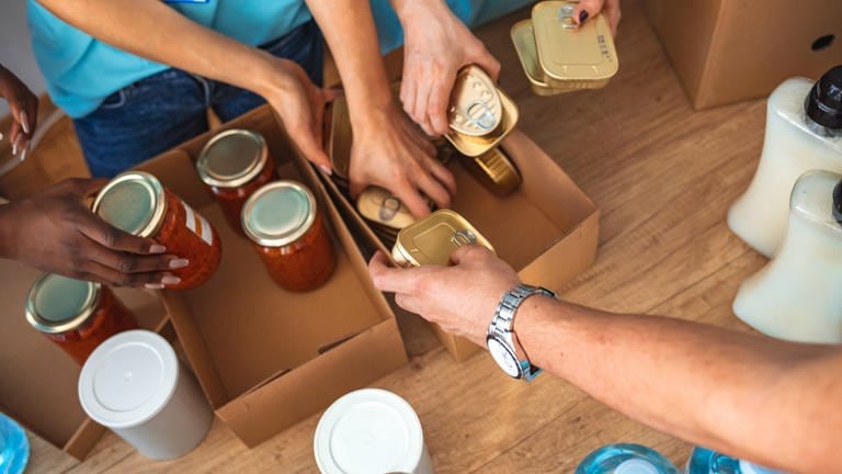 Mehrere Hände Hände greifen nach mit Tomatensoße gefüllten Konservengläsern und goldenen Sardellen-Blechdosen, die in zwei Kartons gestapelt sind.