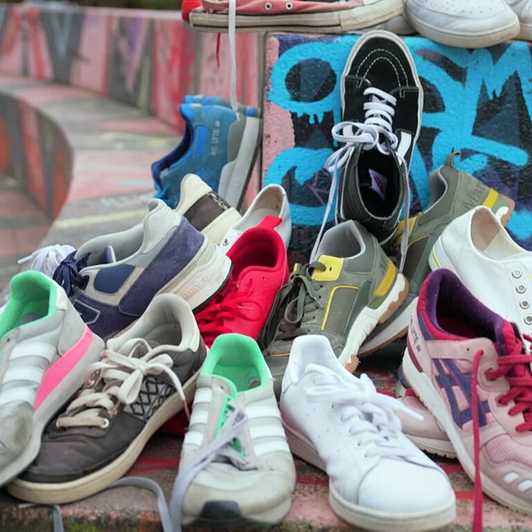 Mehrere Sneaker von Marken wie Vans, Adidas, Nike und Veja liegen auf einem Haufen auf einer mit Graffiti besprayten Mauer.