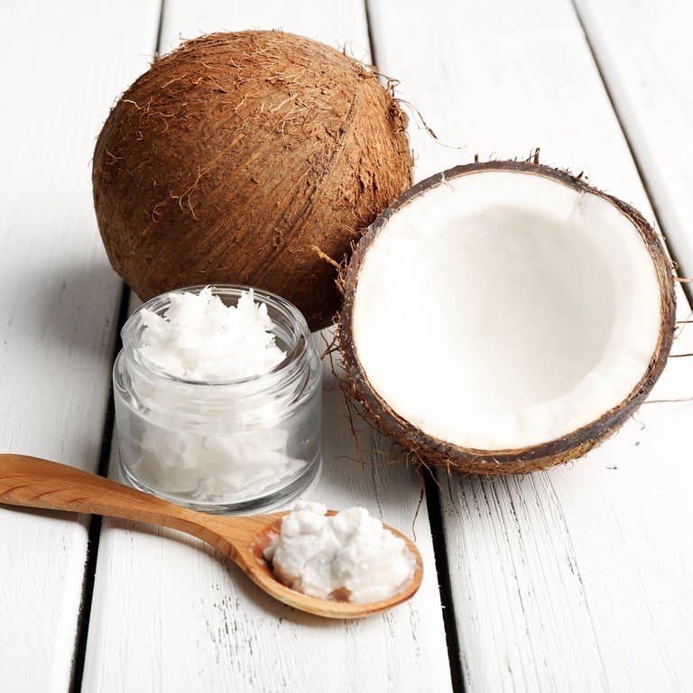 Mittig liegt eine ganze Kokosnuss und eine aufgeschnitte Kokosnuss. Davor steht ein kleiner Glasbehälter mit festes Kokosöl und daneben liegt ein Löffel aus Holz, auf dem das Kokosöl zu schmilzen beginnt.