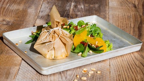 Ein zugeschnürtes Pergamentpäckchen steht neben Salat auf einem Brett (Foto: IMAGO, Imago)