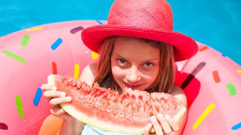 Mädchen liegt in einem aufblasbaren Donut im Pool und isst ein Stück Wassermelone (Foto: IMAGO, Imago)