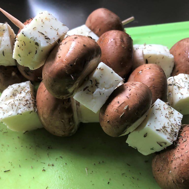 Zwei Champignon-Halloumi-Holzspieße auf einem grünem Schneidebrett. Pilze sind kalorienarm und machen lange satt. Vielfältig variiert sind sie ideal auf dem Grill oder als Antipasti. Gut gewürzt schmecken sie hervorragend als Ersatz für Fleisch.