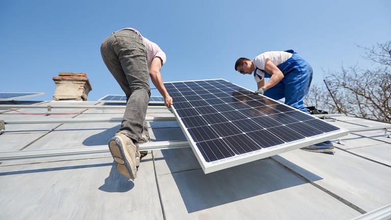 Zwei Männer installieren ein Panel einer Solaranlage auf einem Hausdach. 