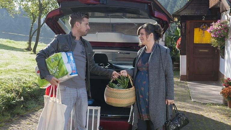 Andreas und die schwangere Eva kommen vom Einkaufen und stehen hinter dem geöffneten Kofferraum ihres Wagens