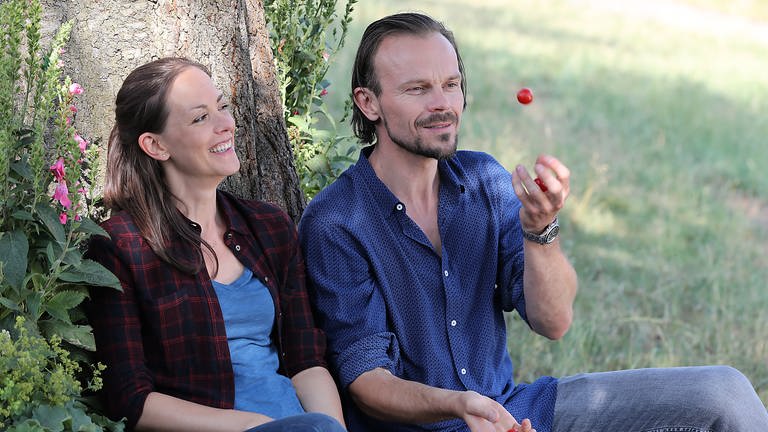 Constantin und Sophie sitzen unter einem Baum, er wirft eine Erdbeere in die Luft
