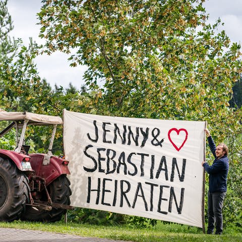Sebastian hält ein riesiges Stück Stoff, das an einem Traktor befestigt ist. Darauf steht "Jenny und Sebastian heiraten" (Foto: SWR, d:light/Christian Koch)