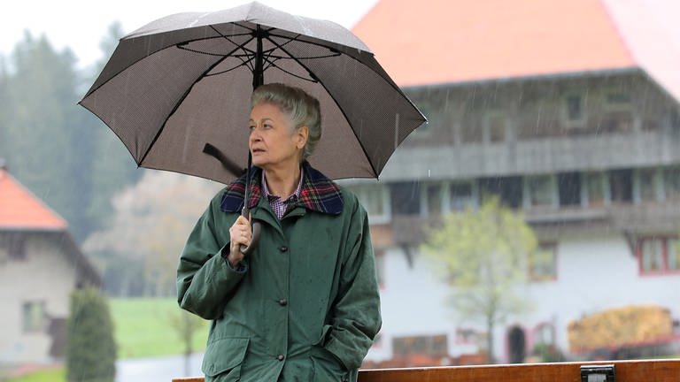 Johanna lehnt mit aufgespanntem Schirm an einer Bank, im Hintergrund der Fallerhof