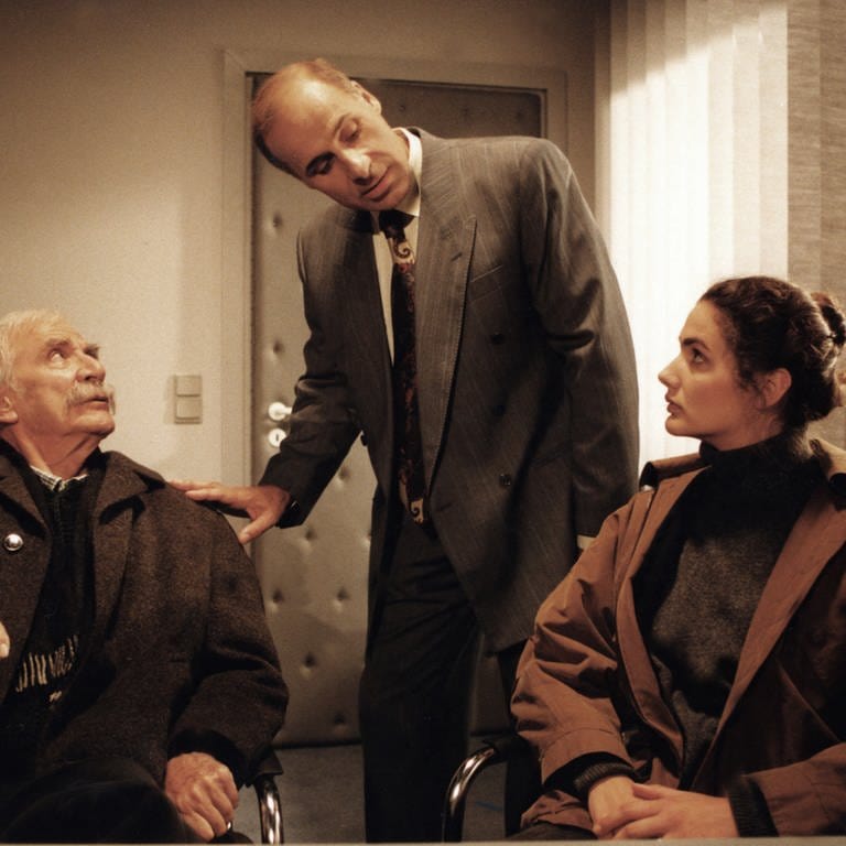 Wilhelm und Monique sitzen, Franz steht dazwischen und legt Wilhelm eine Hand auf die Schulter