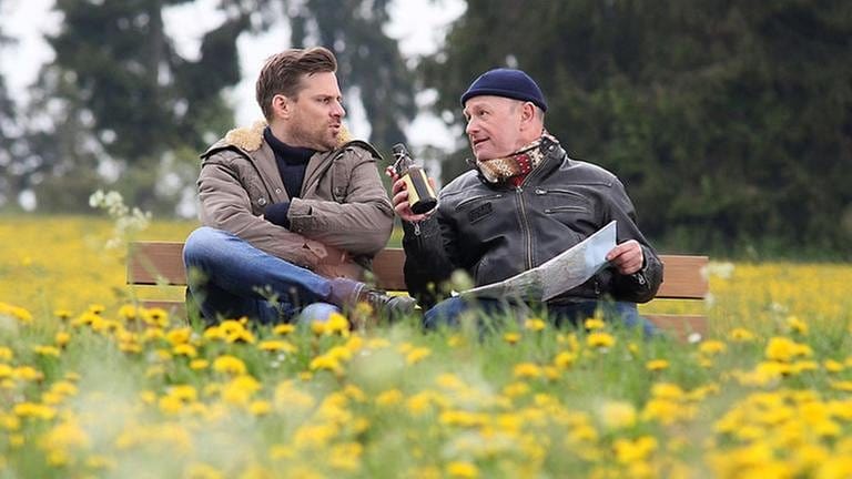 Andreas und Toni sitzen auf einer Bank, Toni hält ein Bier, ringsum blühen Löwenzahnblumen