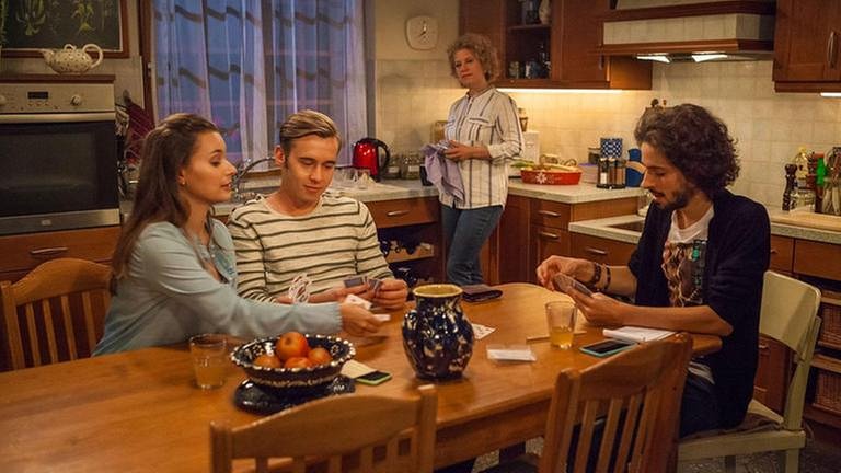 Sebastian, Jenny und Albert spielen am Küchentisch Karten, Bea steht im Hintergrund und trocknet Geschirr ab