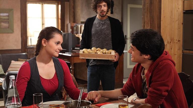 Jenny und Daniel sitzen händchenhaltend am Tisch, dahinter steht Albert, der eine Kartoffelkiste trägt