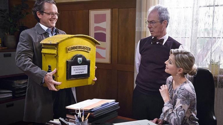 Bernhard bringt einen großen gelben Briefkasten ins Bürgermeisteramt, Frau Heilert und Herr Weiss staunen